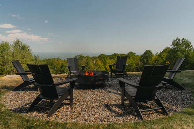 Circular fire pit with glowing flames surrounded by black Adirondack chairs on gravel, overlooking a green forested valley and distant horizon under a clear blue sky.