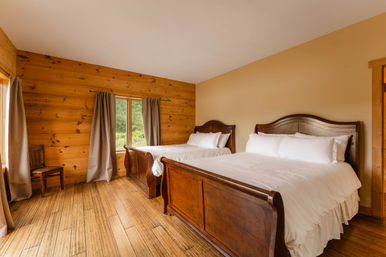 Cozy rustic cabin bedroom with two wooden sleigh beds dressed in white linens, wood-paneled walls and hardwood floors, neutral curtains framing a window with a green forest view.