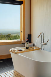 Freestanding white bathtub with wooden tray holding a wine bottle and glass, chrome faucet running, large window framing rolling countryside view