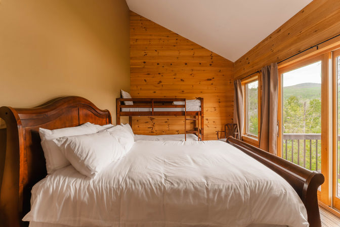 Cozy mountain cabin bedroom with white-sheathed sleigh bed, wooden bunk bed, knotty pine walls and sliding glass doors opening to a forested hillside view.