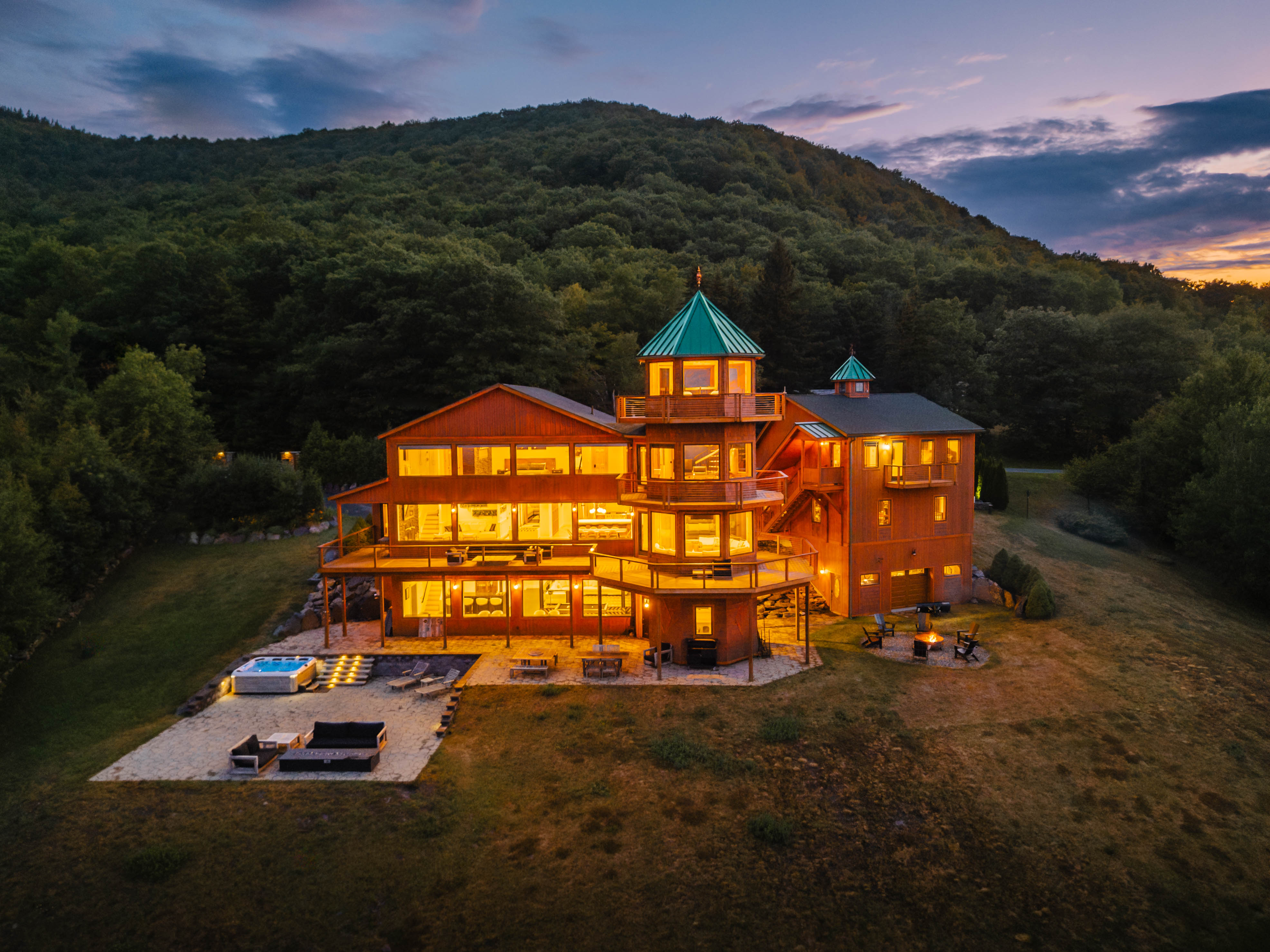 Multi-story illuminated wooden mountain lodge with turret and wraparound decks at sunset, cozy warm glow inside, stone patio with hot tub and outdoor seating on a grassy clearing surrounded by forested hillside.