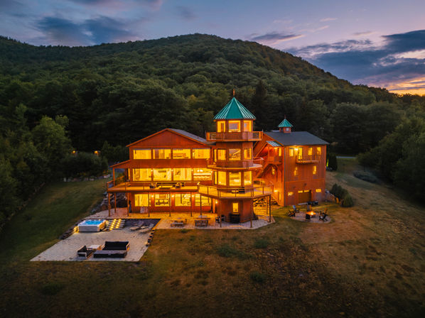 Multi-story illuminated wooden mountain lodge with turret and wraparound decks at sunset, cozy warm glow inside, stone patio with hot tub and outdoor seating on a grassy clearing surrounded by forested hillside.