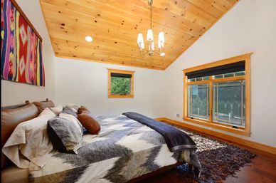 Cozy mountain-style bedroom with angled pine plank ceiling and chandelier, queen bed with gray-and-white geometric duvet and leather pillows, colorful woven wall hanging, hardwood floors, shag rug, and large windows looking out to green trees.