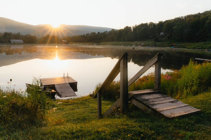 Golden sunrise over a glassy mountain lake with a small wooden dock and weathered steps on a grassy shore, morning mist