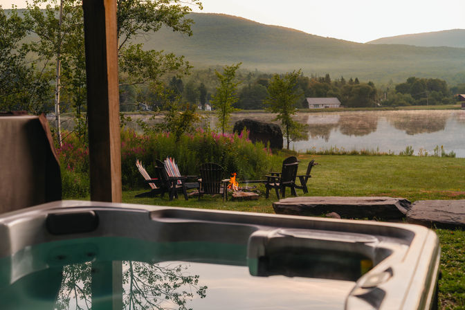 Lakeside mountain retreat at sunrise — hot tub in the foreground, Adirondack chairs around a crackling fire pit, mist rising over a calm lake and rolling green hills.