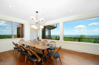 Bright coastal dining room with a long live-edge wooden table for eight, mid-century chairs, a modern chandelier and hardwood floors, framed by large windows with panoramic treeline and ocean views.