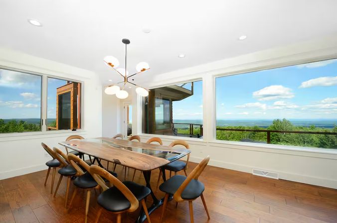 Bright coastal dining room with a long live-edge wooden table for eight, mid-century chairs, a modern chandelier and hardwood floors, framed by large windows with panoramic treeline and ocean views.