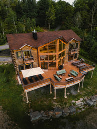 Aerial view of a lakefront log cabin with a large wraparound wooden deck, panoramic floor-to-ceiling windows, outdoor lounge seating and umbrella overlooking rocky shoreline and dense green forest.
