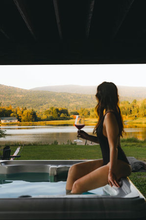 Person in a black swimsuit sitting in a hot tub holding a glass of red wine, overlooking a peaceful lake and tree-covered hills bathed in warm golden-hour light.