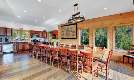Sunny open-concept kitchen and rustic dining room with a long wooden table seating many, wood-paneled walls, hardwood floors, pendant chandelier, and large windows framing a forest view.