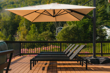Sunlit wooden deck with three black lounge chairs under a beige cantilever umbrella overlooking a pink wildflower meadow and forested hillside.
