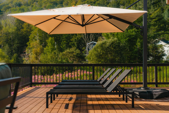 Sunlit wooden deck with three black lounge chairs under a beige cantilever umbrella overlooking a pink wildflower meadow and forested hillside.