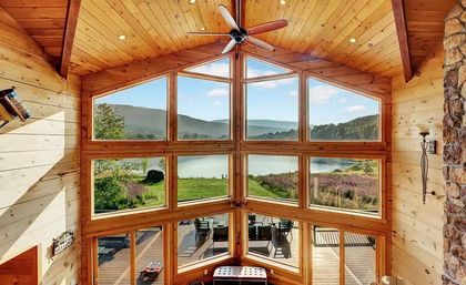 Cozy sunlit log cabin interior with vaulted pine ceiling and fan, floor-to-ceiling panoramic windows framing a lake, rolling hills and a deck with outdoor chairs.