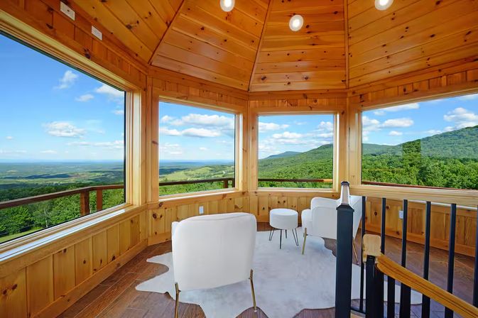Wood-paneled mountain lookout with panoramic windows, two white lounge chairs and ottoman overlooking green ridges and a blue sky