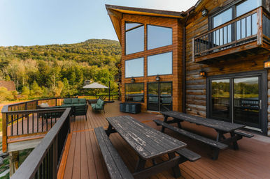 Sunlit mountain cabin deck with wooden picnic tables, cushioned lounge seating, large panoramic windows and lake and forest views.
