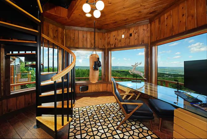 Cozy wood-paneled mountain cabin office with black spiral staircase, hanging punching bag, geometric rug and glass desk with leather chair facing panoramic windows overlooking a green valley and blue sky.