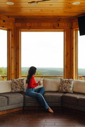 Woman in a red top holding a mug, sitting barefoot on a gray sectional in a wood‑paneled mountain cabin, gazing out large panoramic windows at rolling forested hills.