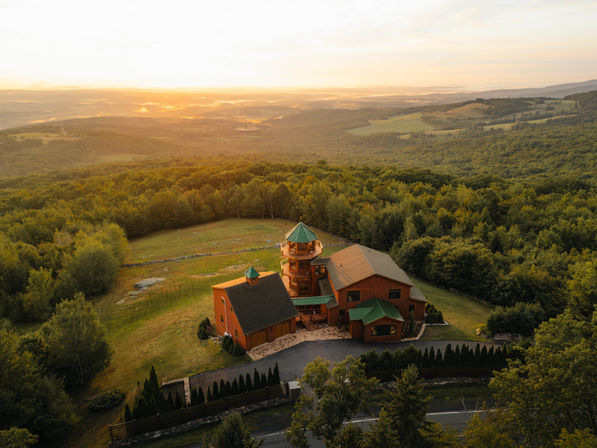 Aerial view of a picturesque wooden mountain lodge with green metal roofs and a round lookout tower on a grassy clearing, surrounded by dense forest and rolling hills bathed in golden sunset light.