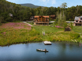 Lakefront log cabin nestled in forested mountains above a wildflower meadow, with a small dock and a person paddling a rowboat on calm blue water.