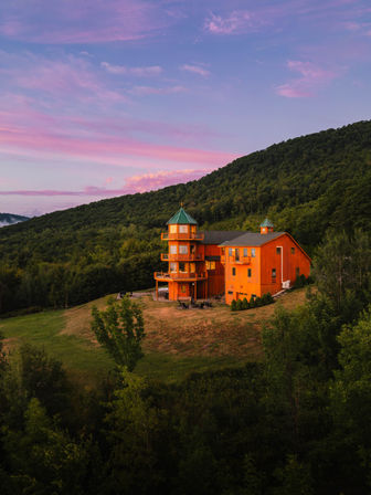Orange wooden mountain lodge with turret and green cupola on a grassy hillside surrounded by forest under a pink and purple sunset sky