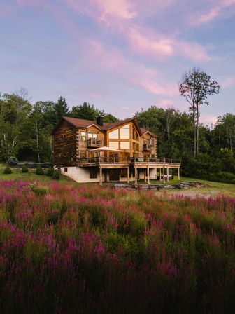 Rustic log cabin with a wide wooden deck overlooking a vibrant purple wildflower meadow and surrounding forest under a pink sunset sky — peaceful countryside retreat