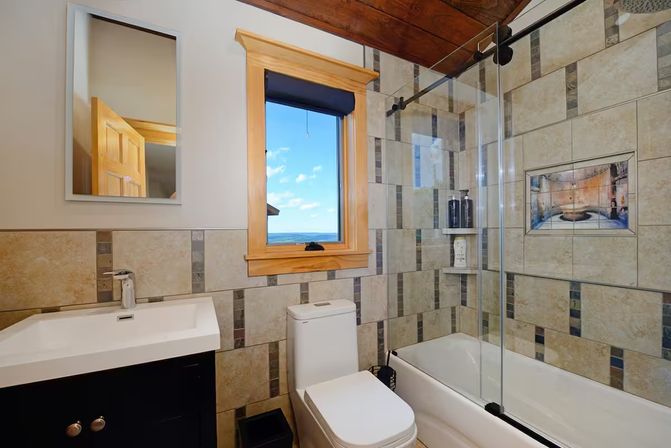 Modern rustic tiled bathroom with white vanity and toilet, bathtub with glass sliding shower door, decorative tile niche, and wood-trim window framing a bright blue ocean view