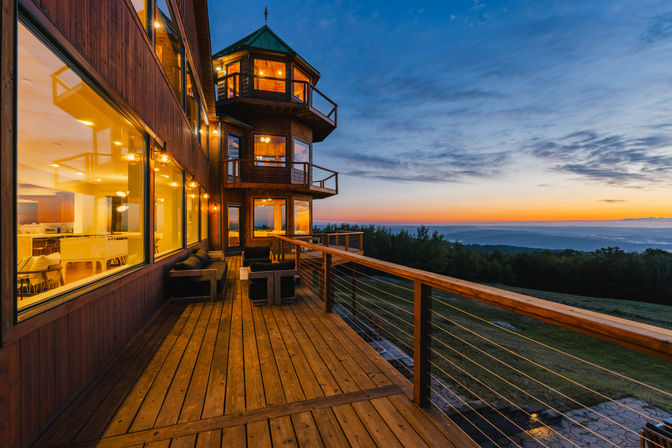 Sunset view from a warm-lit wooden mountain lodge deck with cozy outdoor seating, multi-level tower balconies, large windows and panoramic forested hills