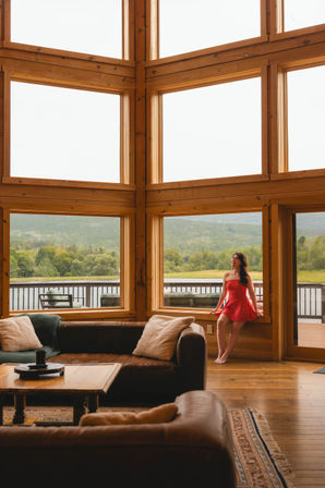 Cozy lakeside cabin living room with floor-to-ceiling wooden windows overlooking a lake and forested hills; a woman in a red polka-dot dress sits on the windowsill with leather sofas and a wooden coffee table in the foreground.