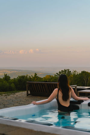 Person relaxing in an outdoor hot tub on a stone patio, overlooking rolling green hills and trees beneath a pastel sunset sky