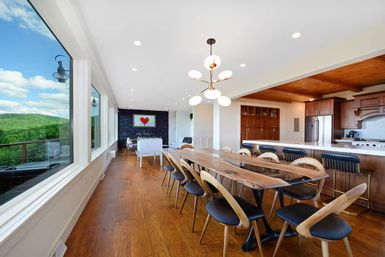 Sunlit open-concept dining room and kitchen with a long live-edge wood table, curved mid-century chairs, brass pendant chandelier, hardwood floors and floor-to-ceiling windows framing panoramic green hill and mountain views.