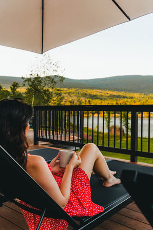 Person in a red sundress lounging on a lakefront deck under an umbrella, holding a mug and enjoying mountain and water views in warm golden light.