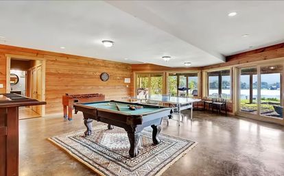 Lakefront game room with wood-paneled walls and polished concrete floor featuring a pool table on a patterned rug, ping-pong table, foosball, dartboard and seating by large glass doors with panoramic lake views.