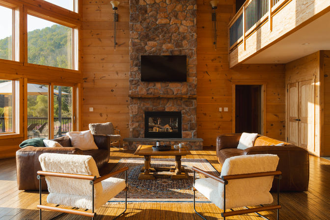 Sunlit mountain-lodge living room with floor-to-ceiling windows, wood-paneled walls, stone fireplace with mounted TV, leather sofas, cozy shearling armchairs, wooden coffee table on a patterned rug and loft balcony.