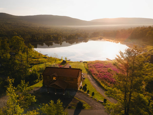 Aerial view of a rustic wooden cabin on a grassy shore beside a calm mountain lake at golden hour, purple wildflower meadow, tree-lined hills and small docks reflecting sunlight.