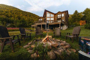 Mountain log cabin retreat with wraparound deck, Adirondack chairs around a glowing fire pit on a grassy meadow and forested hillside backdrop