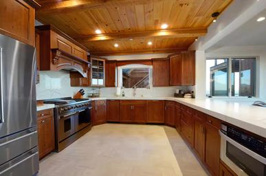 Spacious U-shaped kitchen with warm knotty-wood ceiling and cabinetry, white quartz countertops, stainless steel range and refrigerator, and a sink under a window with an outdoor view.