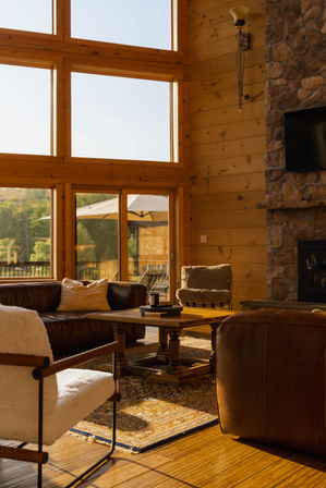 Cozy rustic cabin living room with floor-to-ceiling windows, warm wood-paneled walls, leather sofa and plush chairs around a wooden coffee table on a patterned rug, stone fireplace, and sunlit outdoor deck with umbrella.