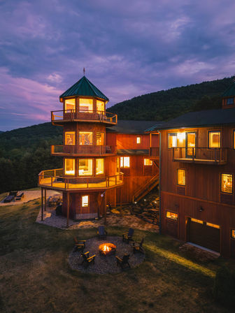 Cozy multi-level wooden mountain lodge with an illuminated round tower, balconies, and a glowing fire pit circle at twilight against forested hills.