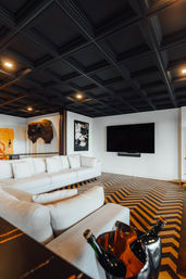 Modern living room with sleek black coffered ceiling, plush white sectional sofa, chevron wood floor, wall-mounted TV, decorative bison head on the wall, and champagne bottles in an ice bucket — stylish media lounge.