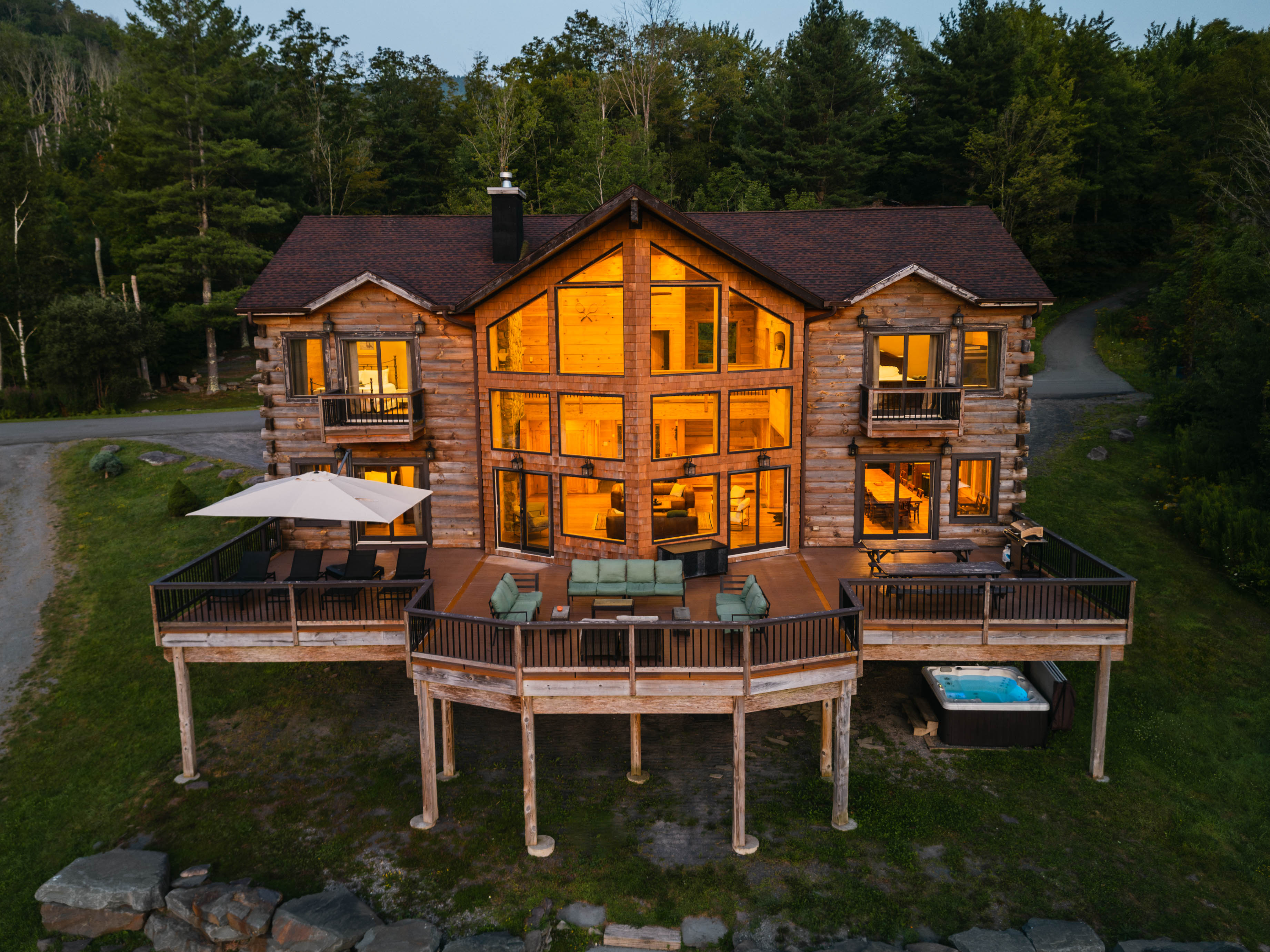 Aerial dusk view of a large rustic log cabin with warm floor-to-ceiling windows, wraparound deck with lounge seating, umbrella and hot tub, set on a wooded hillside beside a country road.