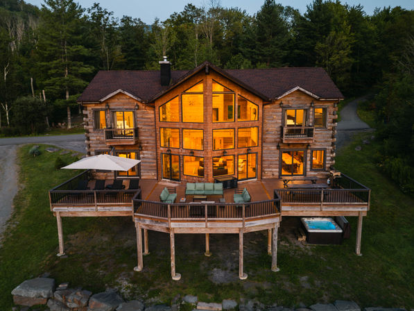 Aerial dusk view of a large rustic log cabin with warm floor-to-ceiling windows, wraparound deck with lounge seating, umbrella and hot tub, set on a wooded hillside beside a country road.