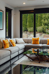 Contemporary living room with a large gray sectional sofa, yellow script-accent pillows, glass coffee table, and floor-to-ceiling windows flooding the space with natural light and a manicured evergreen garden view.