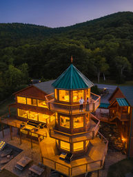 Twilight view of a glowing multi-level wooden tower lodge with a green metal roof and wraparound decks overlooking forested mountains, a couple standing on the upper balcony.