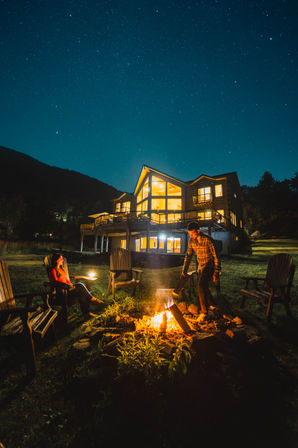 Two people gathered around a glowing campfire by Adirondack chairs in front of a warmly lit mountain cabin under a clear, star-filled night sky