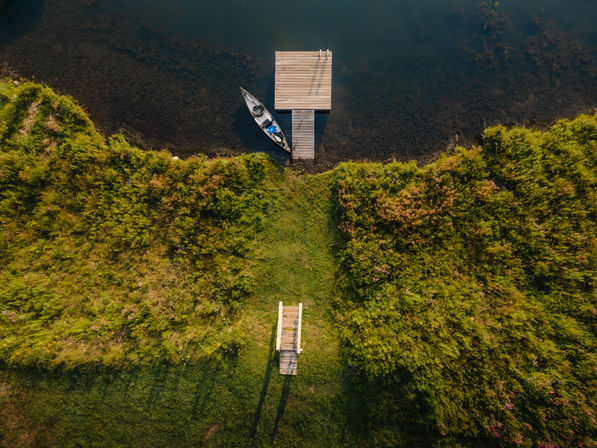 Aerial drone view of a small wooden dock with a canoe tied on calm dark water, flanked by lush green shoreline and a grassy stair path leading down to the pier.