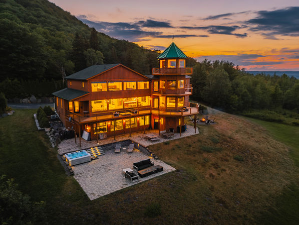 Warm-lit luxury hilltop cabin with green-roofed turret, hot tub and stone patio overlooking forested mountains at sunset
