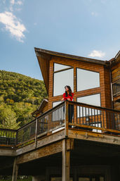 Woman in a red sweater on a rustic wooden cabin balcony, gazing over a green forested mountain slope beneath a clear blue sky.