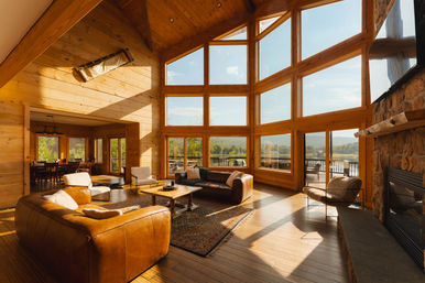 Sunlit rustic great room in a lakefront mountain lodge with wood-paneled walls, floor-to-ceiling windows offering panoramic lake and forest views, leather sofas, stone fireplace, dining area and deck.