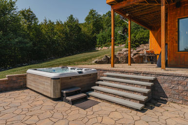 Inviting backyard hot tub and outdoor spa on a patterned stone patio beside a wooden deck with concrete steps, grassy lawn and wooded hillside backdrop