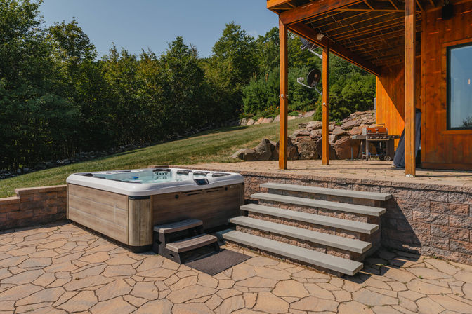 Inviting backyard hot tub and outdoor spa on a patterned stone patio beside a wooden deck with concrete steps, grassy lawn and wooded hillside backdrop